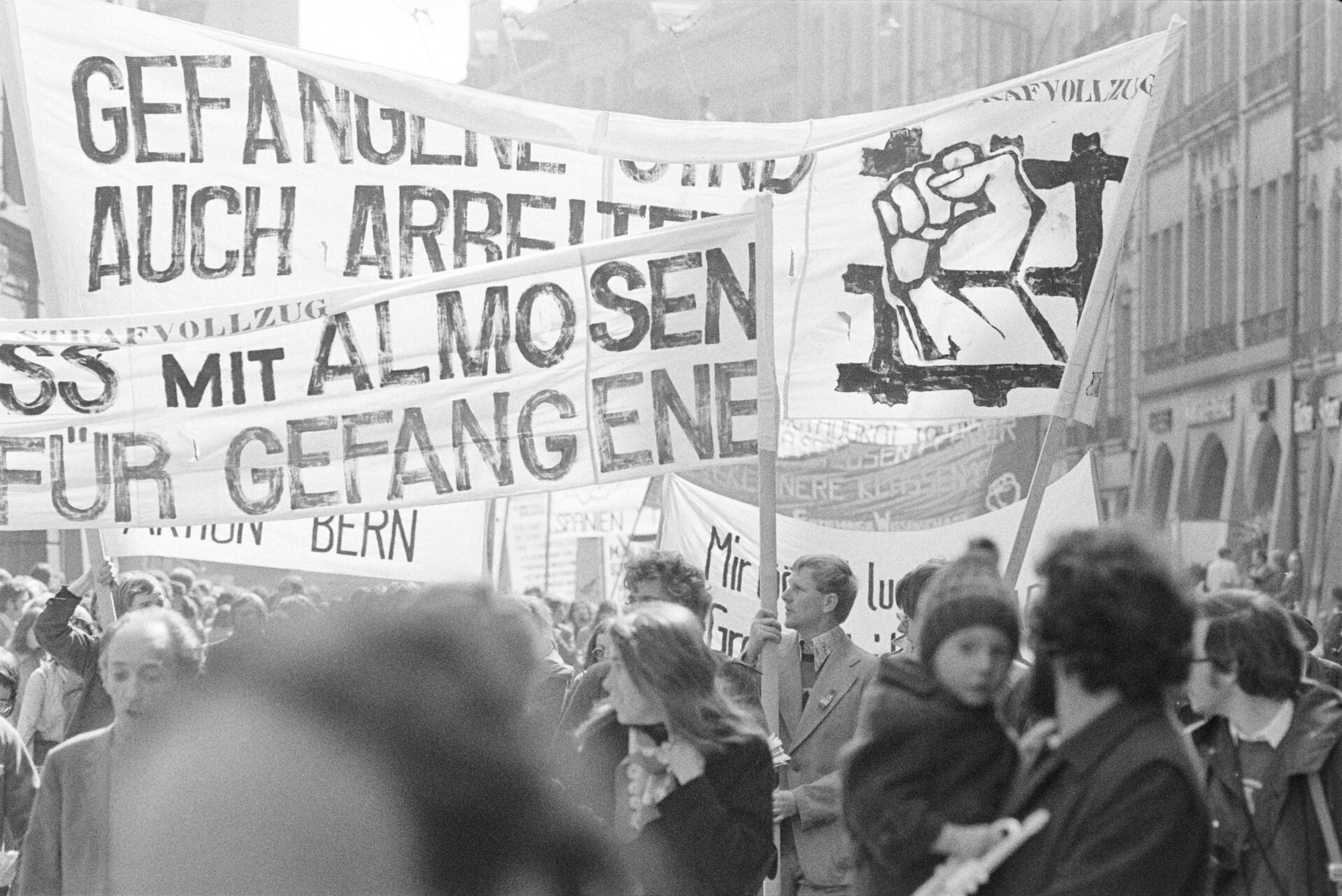 Schwarz-Weiss-Fotografie einer 1. Mai-Demontration in Bern 1976. Slogans auf den Transparenten: "Gefangene sind auch Arbeiter" und "Schluss mit Almosen für Gefangene"