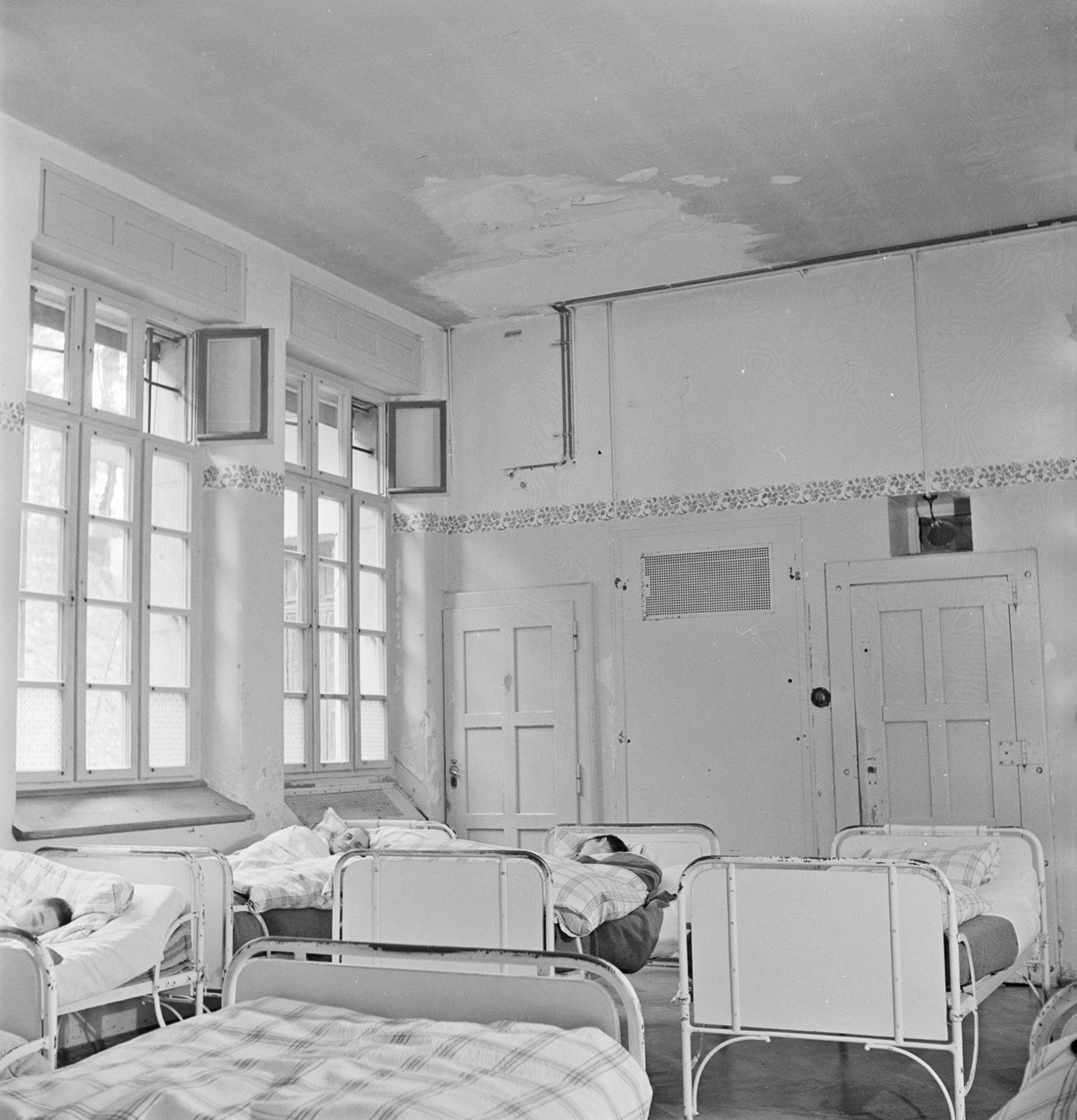 Schwarz-Weiss-Fotografie eines Zimmers in einer psychiatrischen Klinik um 1948. Es sind mehrere Metallbetten zu sehen, in dreien liegt jemand, die Personen haben den Kopf abgewandt.