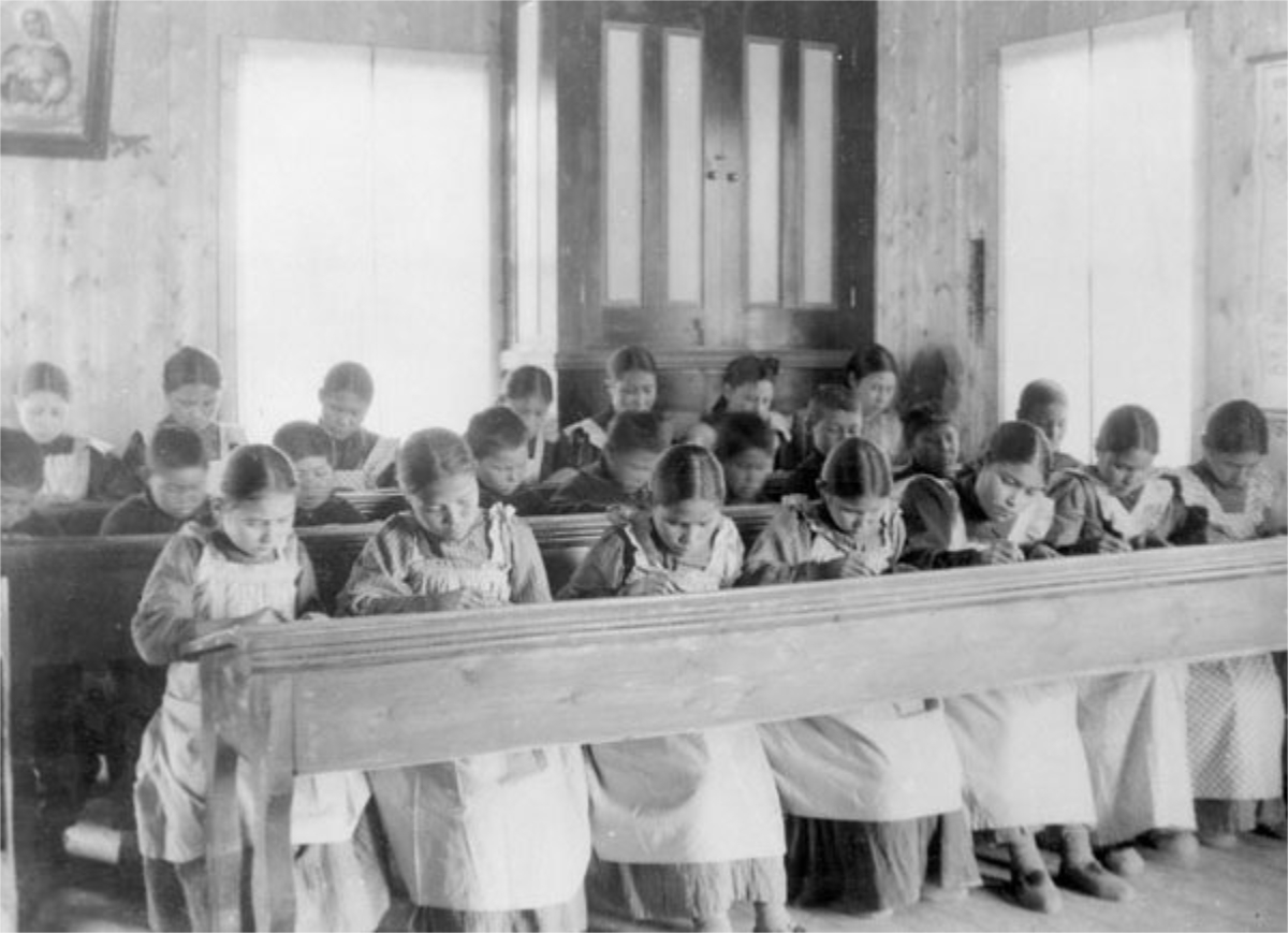 Schwarz-Weiss-Fotografie eines Klassenzimmers mit hölzernen Schulbänken, in welchen junde Mödchen sitzen und schreiben. Es handelt sich um eine sogenannte "Residential School" (Erzieungsheim) für indigene Kinder in Kanada.