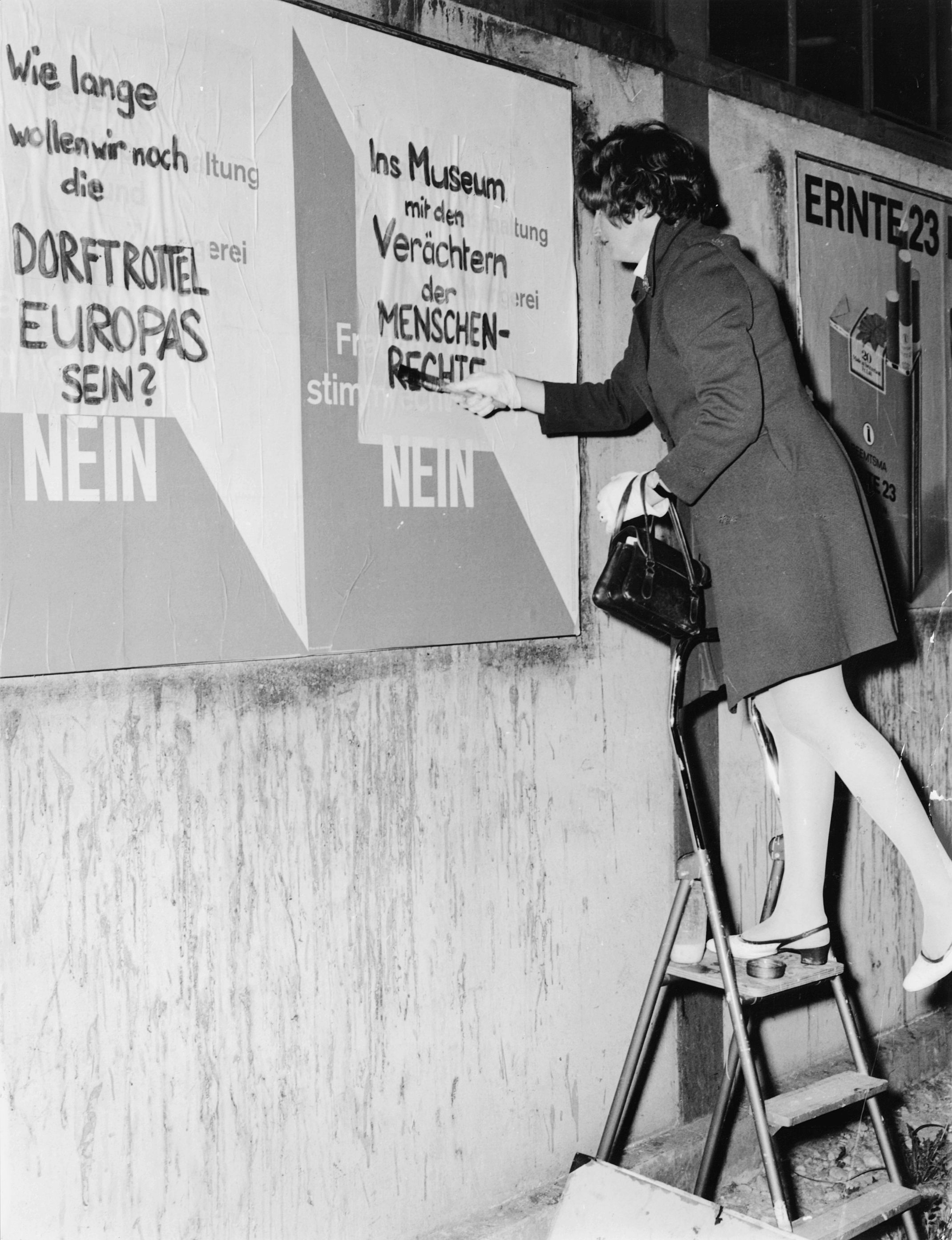 Schwarz-Weiss-Fotografie einer Frau, die auf einer Leiter staht und ein Abstimmungsplakat von Gegnern des Frauenstimmrechts überschreibt. Der Text auf ihren Plakaten lautet: "Wie lange wollen wir noch die Dorftrottel Europas sein?" und "Ins Museum mit den Verächtern der Menschenrechte". Die Fotografie entstand vermutlich im Rahmen des "Marschs auf Bern" 1969.