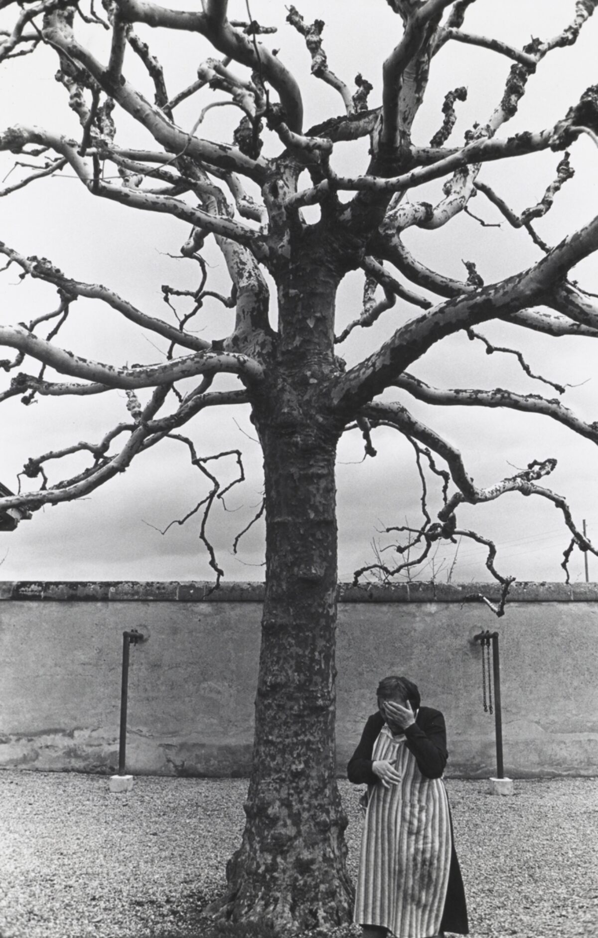 Schwarz-Weiss-Fotografie einer Frau unter einem kahlen Baum. Die Frau hält sich die Hand vor das Gesicht, welches dadurch nicht zu erkennen ist. Das Bild vermittelt Einsamkeit und Hoffnungslosigkeit. Es wurde 1971 in der Psychiatrischen Universitätsklinik Münsingen aufgenommen.