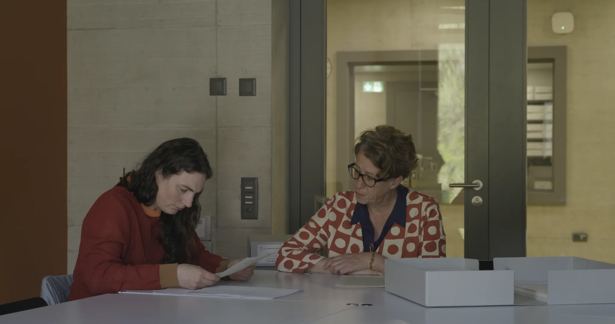 An einem Tisch im Staatsarchiv Zürich sitzt eine Archivarin mit einer Archiv-Besucherin am Tisch. Die Besucherin liest in einer Akte, die Archivarin sitzt daneben.