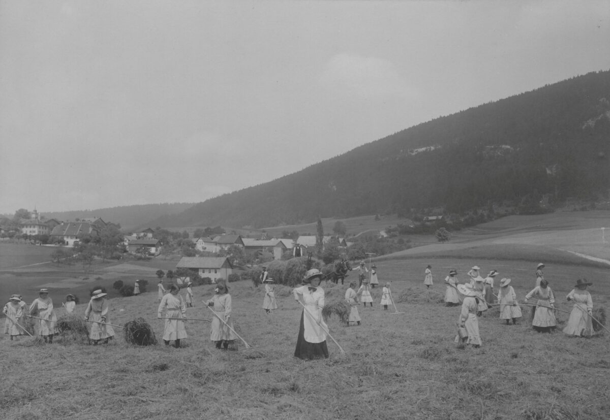 Schwarz-Weiss-Fotografie junger Frauen in Sonntagskleidung bei der Feldarbeit im Erziehungsheim Loveresse. Das Bild stammt aus Band 2 der Fotoalben der Direktion des Armenwesens des Kantons Bern und wurde an der Landesausstellung 1914 in Bern ausgestellt.